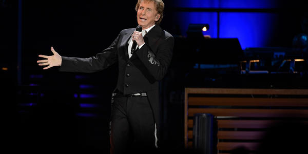 Barry Manilow, wearing a black tailed tuxedo coat, sings into microphone on stage in front of a blue stage lighting.